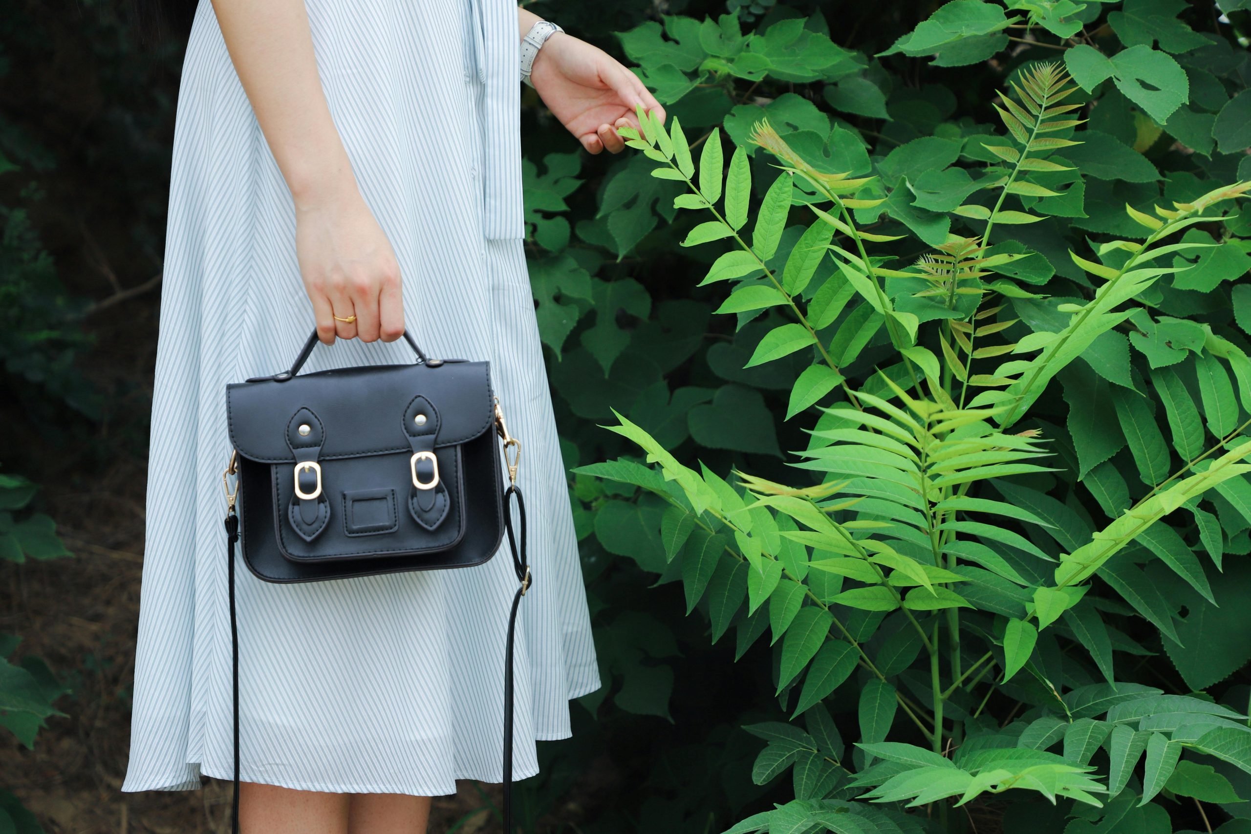 Close-up of a woman holding a black handbag, wearing a striped dress, beside lush green plants.